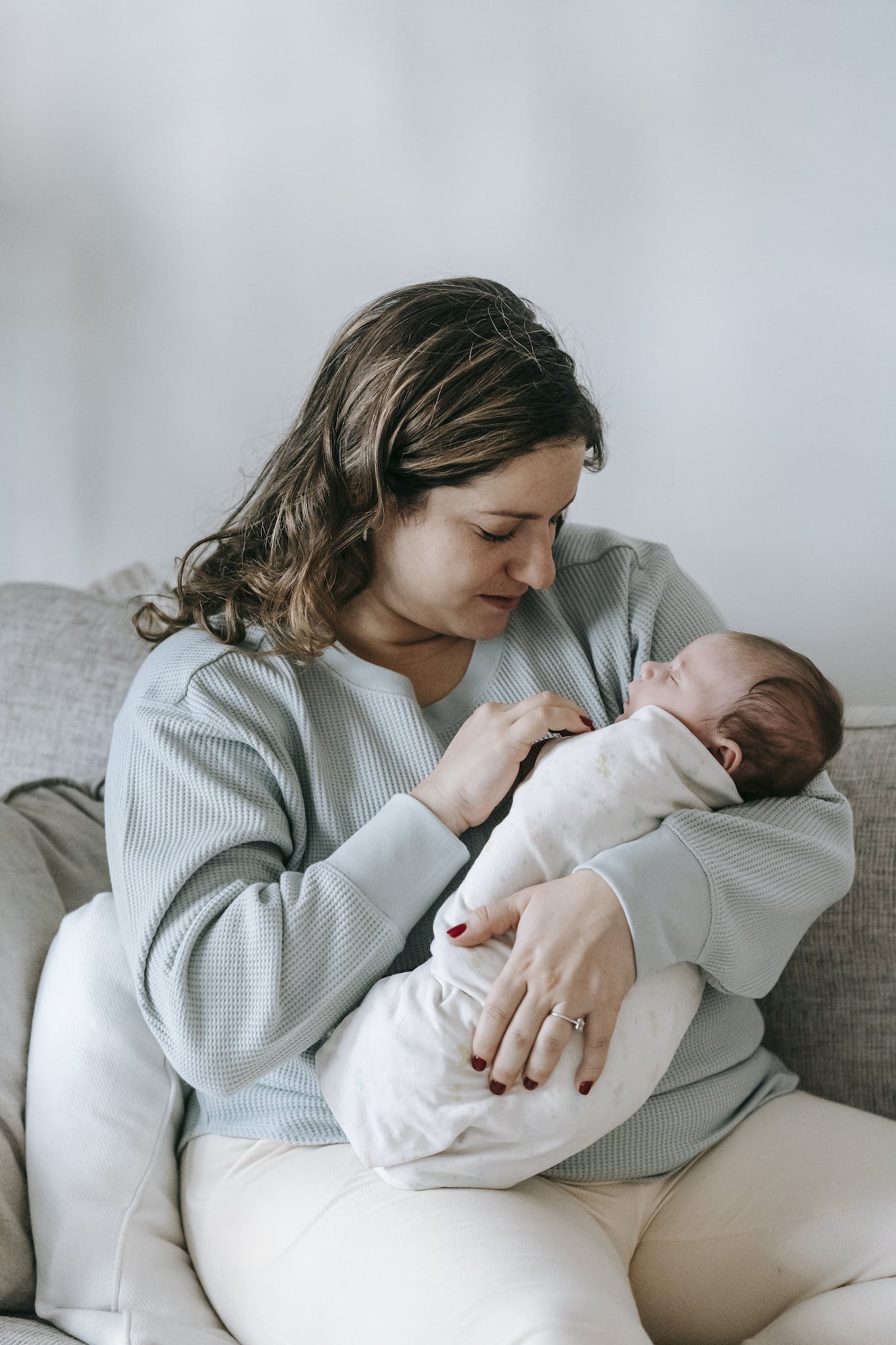 Calm young woman in casual clothes caressing and embracing adorable sleeping baby while sitting on comfortable couch at home