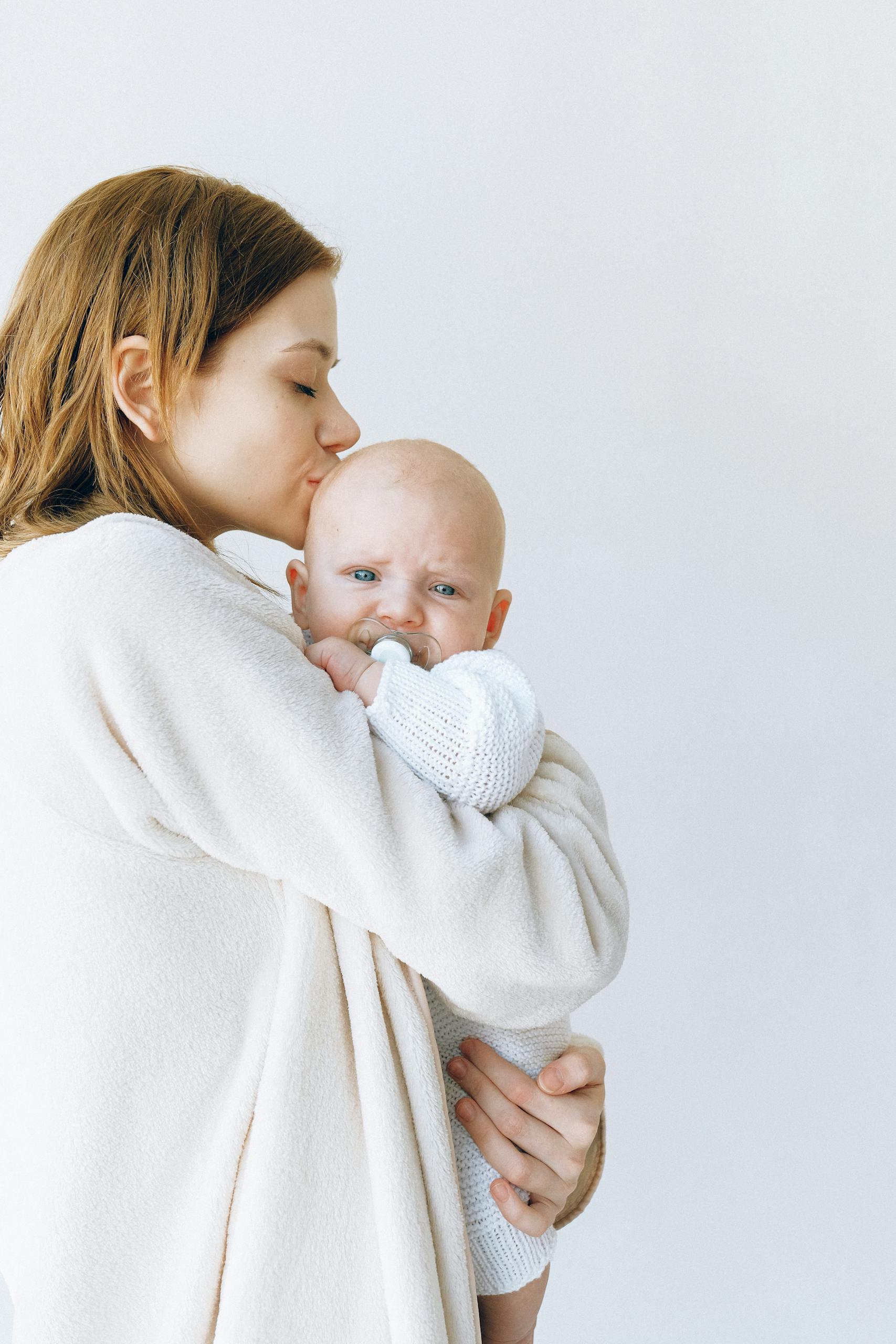 Loving mother gently kisses her calm baby wrapped in a warm sweater.